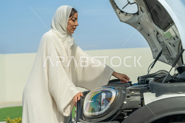 محاولة اكتشاف سبب العطل، الحيرة وعدم معرفة سبب المشكلة, اعتماد المرأة على نفسها في مواجهة مشكلات السيارة، إمرأه عربية خليجية إماراتيه محجبة تتفحص سيارتها بإيماءات القلق، حرية القيادة للمرأة الخليجية، شابة سعودية ترتدي العباءة, الاستقلال عند المرأة العربية