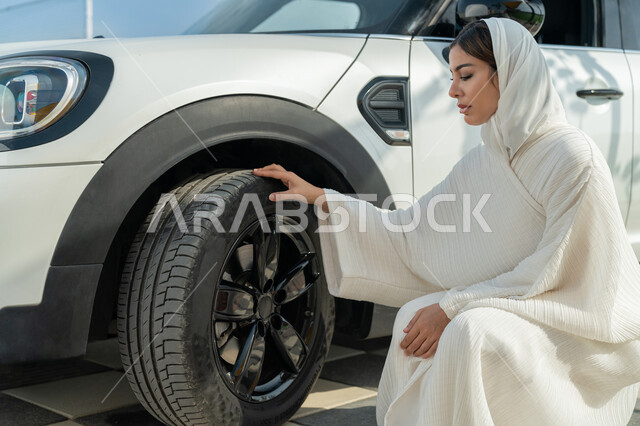 مفهوم الاستقلال والاعتماد على النفس، اعتماد المرأة على نفسها في التنقل و المواصلات، إمرأه عربية خليجية إماراتيه محجبة تجلس بجانب سيارتها، حرية القيادة للمرأة العربية، سيده سعودية ترتدي العباءة تقوم بالتأكد من إطارات السيارة