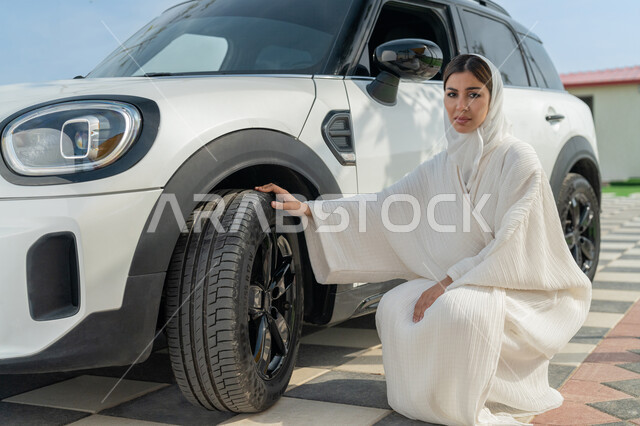 اعتماد المرأة على نفسها في التنقل و المواصلات، مفهوم الاستقلال والاعتماد على النفس، إمرأه عربية خليجية إماراتيه محجبة تجلس بجانب سيارتها، حرية القيادة للمرأة العربية، سيده سعودية ترتدي العباءة تقوم بالتأكد من إطارات السيارة