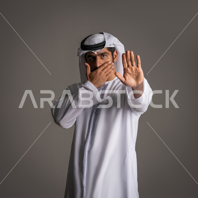 Standing angrily and rejecting a specific matter, silence and stopping talking, a portrait of a young Arab Gulf Emirati man wearing a kandora and ghanara, raising his palm up with gestures of annoyance and covering part of his face, requesting non-interference and refraining from doing a specific matter, gray background