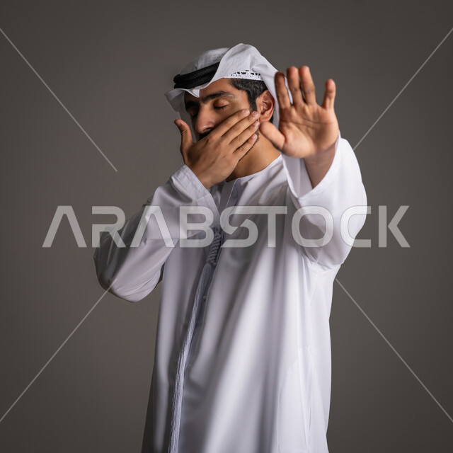 A request not to interfere and refrain from doing a specific thing, standing angrily and rejecting a specific thing, silence and stopping talking, a portrait of a young Arab Gulf Emirati man wearing a kandora and ghanara, raising his palm up with gestures of annoyance and covering part of his face, gray background.