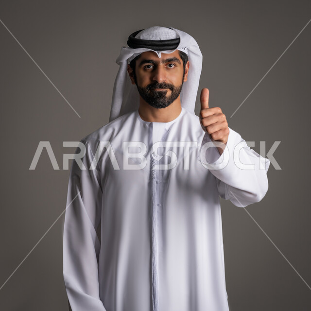 The concept of contentment and admiration, elegance, masculinity, and concern for external appearance, portrait of an Emirati Gulf Arab man wearing a white kandora and ghutra, raising his thumb in a gesture of approval and acceptance, standing upright and looking at the camera with happy expressions, gray background