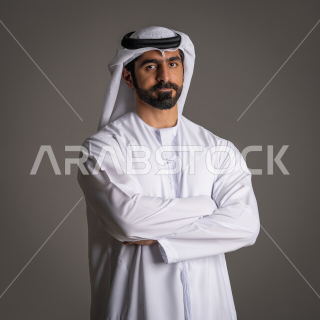 Self-confidence and standing with a straight back, elegance, masculinity and attention to external appearance, portrait of an Arab Gulf Emirati man wearing a white kandora and ghutra and looking at the camera with a cheerful expression, gray background