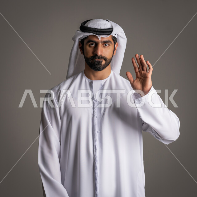 ايماءات إلقاء التحية والسلام والسعادة بالترحيب، بورتريه لرجل عربي خليجي اماراتي يرتدي الكندورة والغترة الاماراتية يقوم برفع يده للأعلى وينظر للكاميرا ببشاشة،  خلفية رمادية
