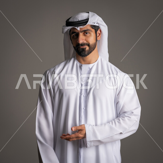 ايماءات الترحيب بسعادة وحب، بورتريه لرجل عربي خليجي اماراتي يرتدي الكندورة والغترة الاماراتية يقوم بفتح كفه ويشير إلى اتجاه معين وينظر للكاميرا ببشاشة،  خلفية رمادية