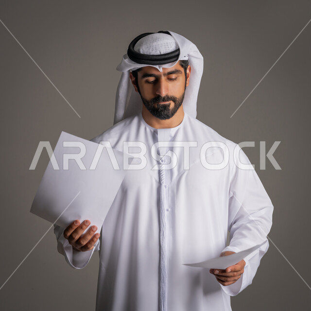 أوراق بيضاء فارغة، استخدام لوحات توضيحية والنظر إليها مفهوم الإعلان والتسويق، موك اب أبيض، بورتريه مقرب لرجل عربي خليجي إماراتي يرتدي الكندورة والغترة يحمل بيده ورقتان فارغتان وينظر إليهما، خلفية رمادية