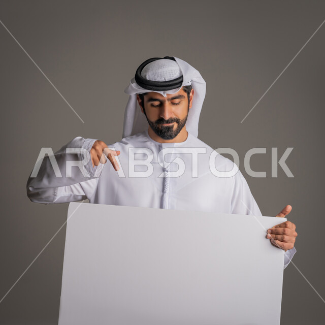 استخدام لوحات توضيحية والإشارة اليها، مفهوم الإعلان والتسويق، لوح إعلاني فارغ، موك اب أبيض، بورتريه مقرب لرجل عربي خليجي إماراتي مبتسم يرتدي الكندورة والغترة يحمل بيده لوحه بيضاء مستطيلة كبيرة وينظر إليها، خلفية رمادية