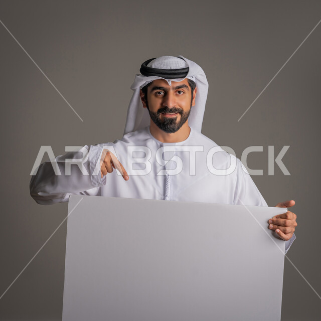 لوح إعلاني فارغ، استخدام لوحات توضيحية والإشارة اليها، مفهوم الإعلان والتسويق، موك اب أبيض، بورتريه مقرب لرجل عربي خليجي إماراتي مبتسم يرتدي الكندورة والغترة يحمل بيده لوحه بيضاء مستطيلة كبيرة وينظر للكاميرا، خلفية رمادية