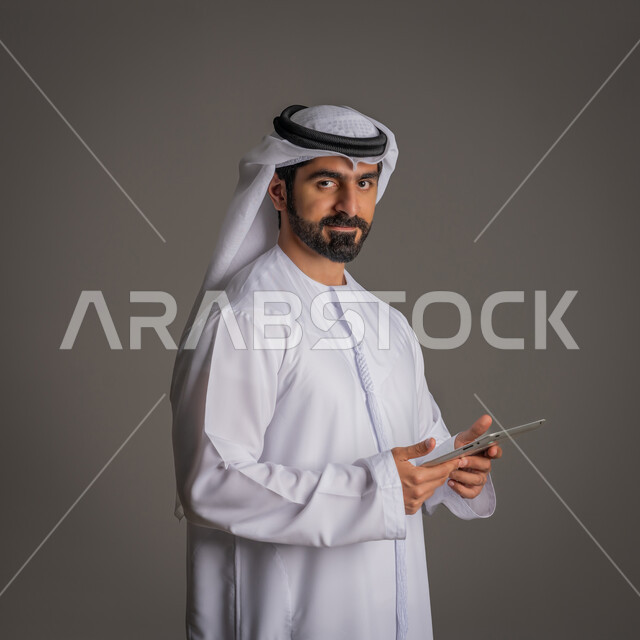 النظر إلى الكاميرا بإيماءات البشاشة, استخدام التكنولوجيا المتطورة للتسويق الرقمي الالكتروني، بورتريه لشاب عربي خليجي اماراتي يمسك الجهاز اللوحي بيده، متابعة أخبار العالم, تصفح الانترنت والصفحات, خلفية رمادية