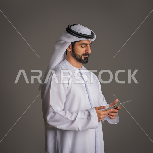 استخدام التكنولوجيا المتطورة للتسويق الرقمي الالكتروني، بورتريه لشاب عربي خليجي اماراتي يمسك الجهاز اللوحي بيده وينظر إليه بإيماءات البشاشة, متابعة أخبار العالم, تصفح الانترنت والصفحات, خلفية رمادية