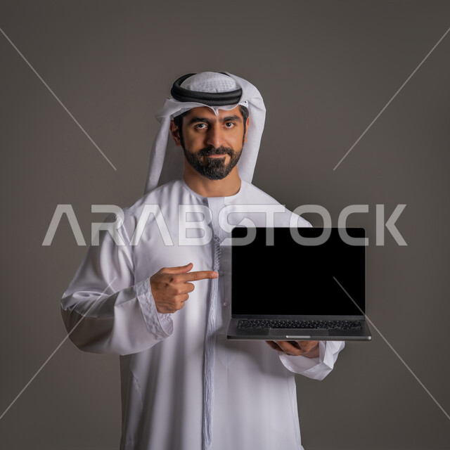 الإشارة باصبع السبابه إلى شاشة سوداء فارغة عبر الحاسوب المحمول، استخدام التكنولوجيا المتطورة للتسويق الرقمي الالكتروني، عرض التطبيقات الحديثة، بورتريه لشاب عربي خليجي اماراتي يمسك اللابتوب بيده وينظر للكاميرا, خلفية رمادية
