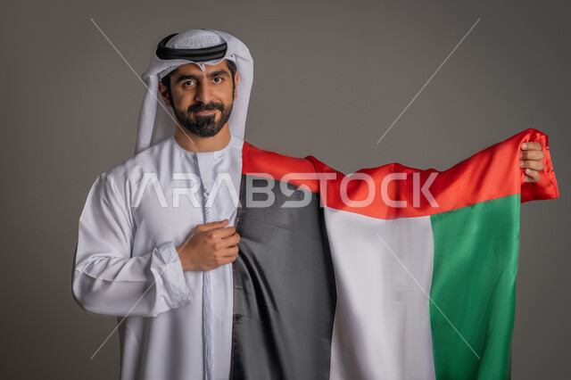إحياء ذكرى الإستقلال لدولة الإمارات العربية المتحدة، حب الوطن والعلم، بورتريه لرجل عربي خليجي إماراتي يرتدي الغترة والكندورة الإماراتية يحمل علم الإمارات، الإحتفال بالعيد الوطني الإماراتي، خلفية رمادية