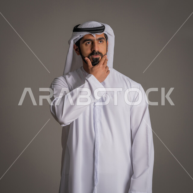 الحيرة والتفكير بشيء ما, بورتريه لشاب عربي خليجي اماراتي يرتدي الكندورة والغترة الاماراتية يقف ويضع يده على ذقنه وينظر لمكان بعيد بإيماءات القلق, التخطيط ووضع الأهداف المستقبلية, خلفية رمادية