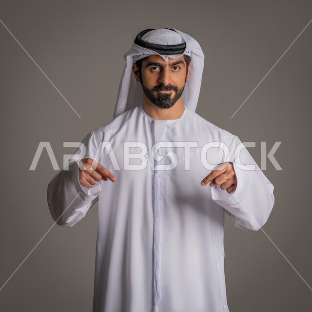 تحديد موقع معين, اشارات تحدد هذا المكان (هنا)، حركات وتعابير وجود شيء ما، بورتريه لرجل عربي خليجي اماراتي يرتدي الغترة والكندورة يشير بأصابعه نحو الأسفل, النظر إلى الكاميرا بإيماءات السرور، خلفية رمادية