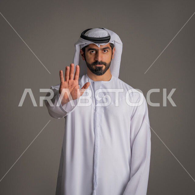 طلب التوقف عن فعل أمر معين وعدم التدخل, الوقوف بغضب ورفض أمر محدد, الامتناع عن فعل شيء ما, بوتريه لشاب عربي خليجي إماراتي يرتدي الكندورة والغنرة يرفع كفه للأعلى بإيماءات الانزعاج, خلفية رمادية