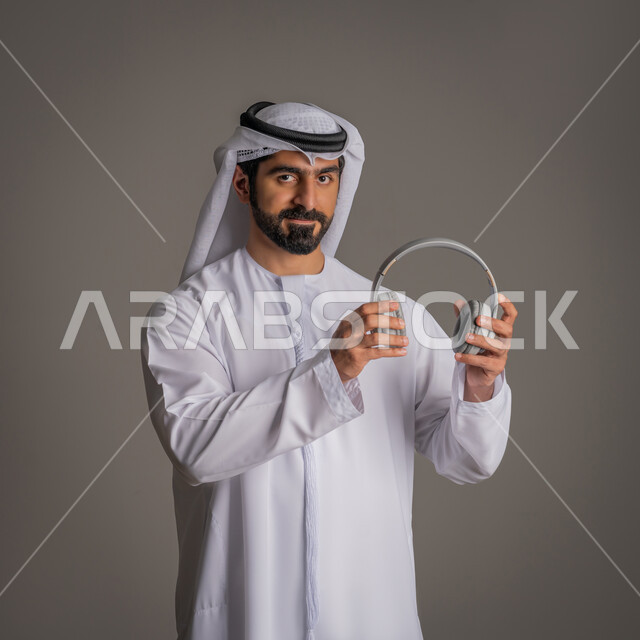 سماعة الاتصال والاستماع للموسيقا, استخدام تقنية الهيدفون, التواصل مع الأهل والأصدقاء, بورتريه لرجل عربي خليجي اماراتي يمسك بيده سماعات الأذن اللاسلكية, تقنيات حديثة وتكنولوجيا, خلفية رمادية