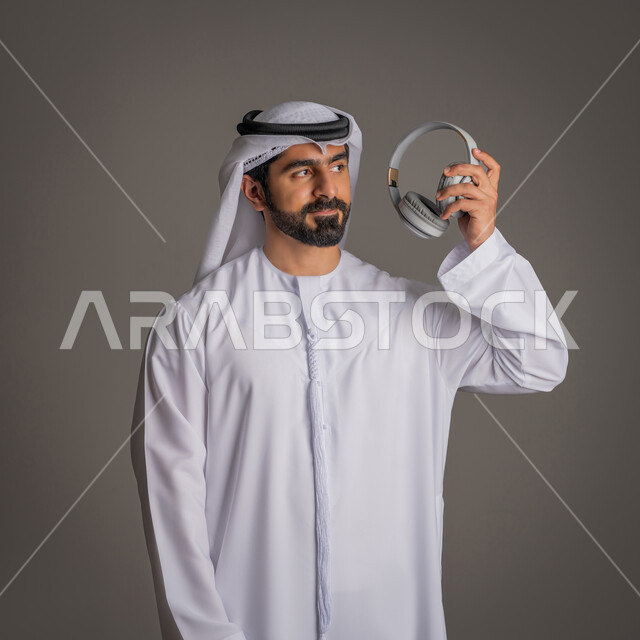 استخدام تقنية الهيدفون, سماعة الاتصال وسماع الموسيقا, التواصل مع الأهل والأصدقاء, بورتريه لرجل عربي خليجي اماراتي يمسك بيده سماعات الأذن اللاسلكية, تقنيات حديثة وتكنولوجيا, خلفية رمادية