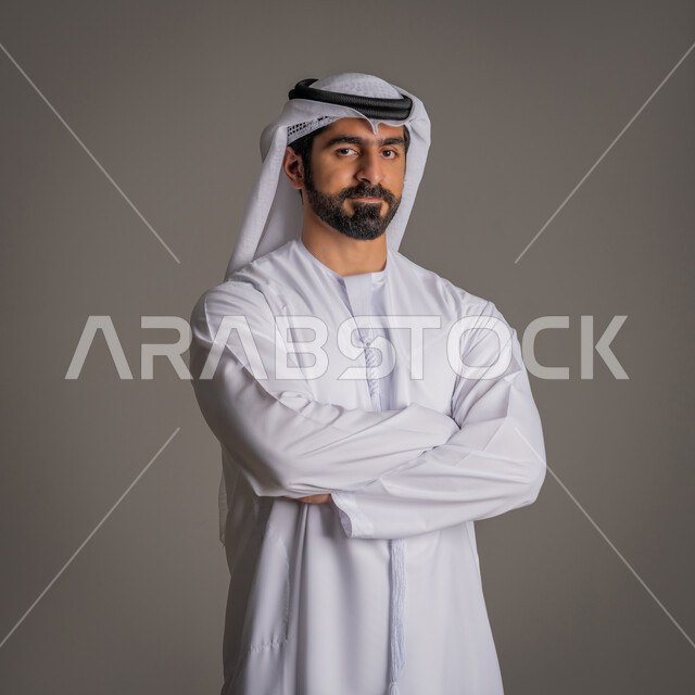 مفهوم الثقة بالنفس والوقوف بظهر مشدود, الأناقة والرجولة والاهتمام بالمظهر الخارجي, بورتريه لرجل عربي خليجي إماراتي يرتدي الكندورة البيضاء والغترة وينظر إلى الكاميرا بتعابير البشاشة, خلفية رمادية