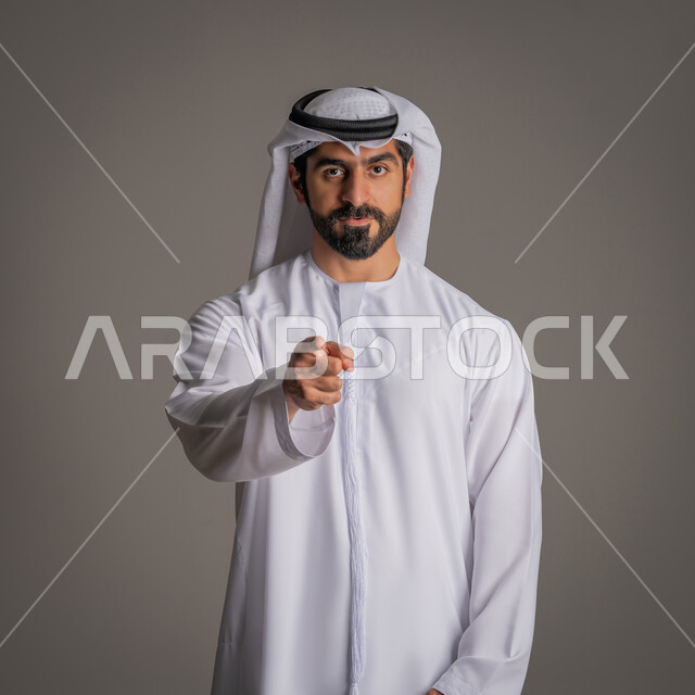 التهديد والتوعد لشيء ما،  الوقوف باستقامة والنظر إلى الكاميرا بإيماءات الغضب, بورتريه لرجل عربي خليجي إماراتي يرتدي الكندورة الاماراتيه والغترة يشير باصبع السبابة للأمام بالاتجاه المقابل, المواجهة والمنافسة, خلفية رمادية