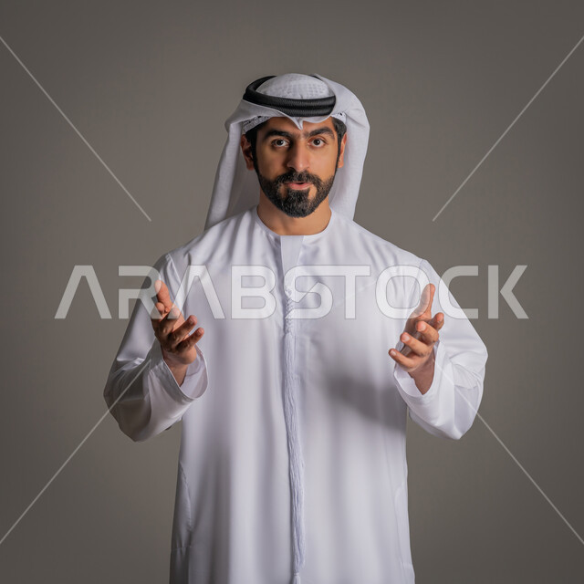 التساؤل والاستفهام عن امر معين, بورتريه لرجل عربي خليجي إمارتي يرتدي الكندورة والغترة الاماراتية يفتح كفيه وينظر الى الكاميرا بإيماءات عدم المعرفة, الاستغراب من حدث ما, خلفية رمادية
