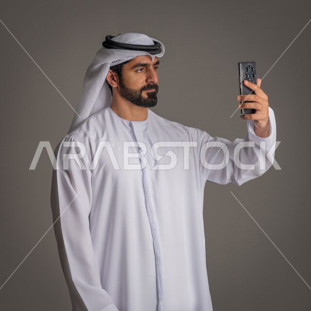 Taking souvenir photos and documenting happy moments, a portrait of an Emirati Gulf Arab man wearing a white kandora and ghutra, holding a mobile phone and taking a selfie of himself, gestures of self-confidence, the concept of elegance, masculinity, and concern for external appearance, gray background