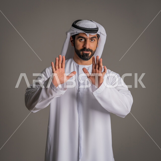 Requesting non-interference, lowering the voice and threats, standing angrily and rejecting a specific order, silence and stopping talking, refraining from doing a specific order, a portrait of a young Arab Gulf Emirati man wearing a kandora and ghanara, raising his palms up with gestures of annoyance, gray background.