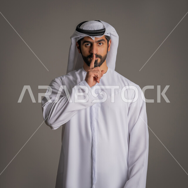 إيماءات طلب السكوت والتوقف عن الكلام، بورتريه لرجل عربي خليجي إماراتي يرتدي الكندورة والغترة ينظر إلى الكاميرا بإيماءات الجدية, وضع اصبع السبابة أمام الفم يشير إلى ضرورة الصمت والتزام الهدوء, خفض الصوت والحاجة إلى السكون, خلفية رمادية