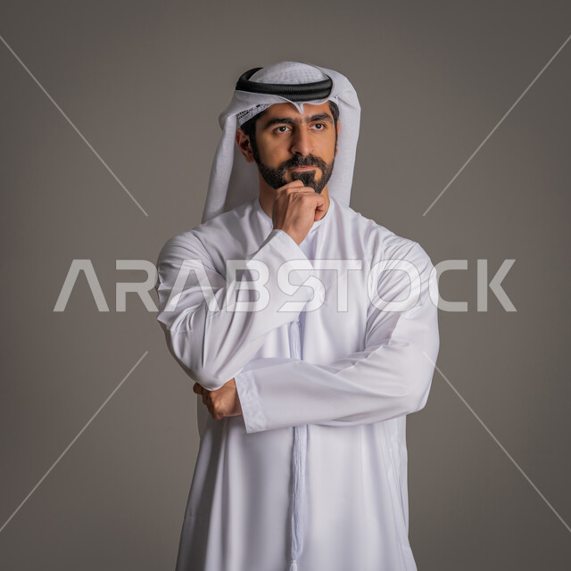 النظر إلى مكان بعيد بشرود, بورتريه لشاب عربي خليجي اماراتي يرتدي الكندورة والغترة الاماراتية يقف ويضع يده أسفل ذقنه بإيماءات القلق والتفكير بشيء ما, التخطيط للمستقبل ووضع الأهداف المستقبلية, خلفية رمادية