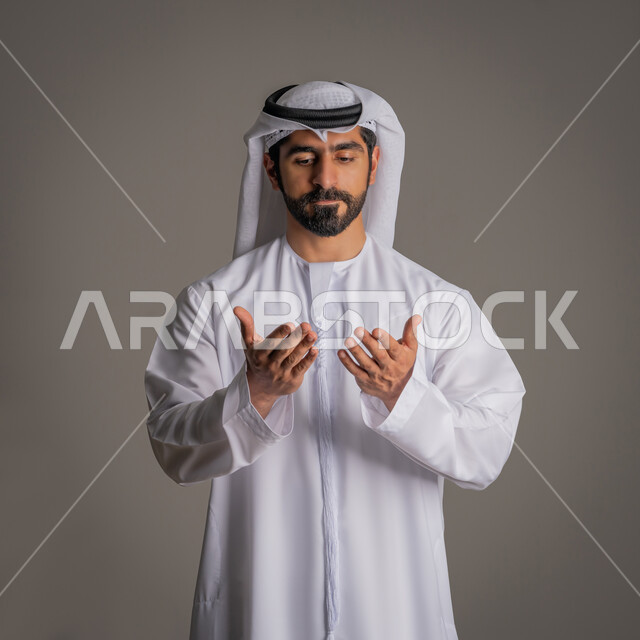 رفع اليدين للدعاء والتقرب إلى الله, بورتريه لشاب عربي خليجي اماراتي يرتدي الكندورة والغترة الاماراتية يقف ويرفع كفيه إلى الأعلى بإبماءات التوسل والتضرع, الاجتهاد في العبادة, خلفية رمادية
