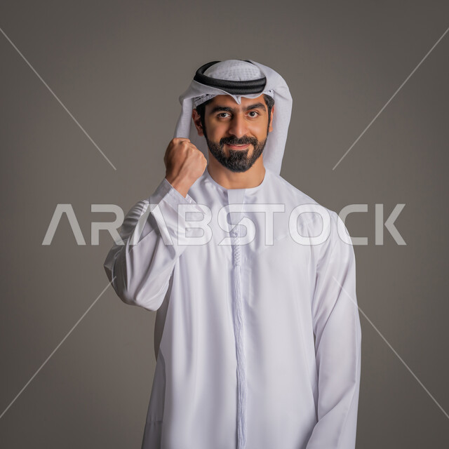 إيماءات الوجه واليدين تدل على الفرح بالنجاح والفوز, بورتريه لرجل عربي خليجي إماراتي مبتسم يرتدي الكندورة الاماراتيه والغترة يرفع قبضة يده للأعلى, الاحتفال بتحقيق الأهداف والانجاز, تعبيرات النصر والانتصار , خلفية رمادية