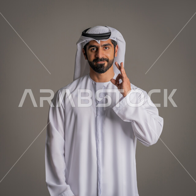 التعبير عن الجودة والاتقان، بورتريه لشاب عربي خليجي إماراتي يرتدي الكندورة الإماراتية والغترة ويشير بيده بإيماءات وتعابير وجه و يدين تدل على الاعجاب، علامة الامتياز والتميز، القبول والموافقة والرضا، خلفية رمادية