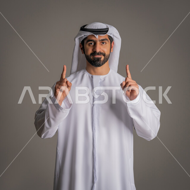 النظر إلى الكاميرا بشكل مباشر بتعابير البشاشة, الوقوف ورفع اصبعي السبابة في كلتا اليدين بإيماءات مختلفة, بورتريه لرجل عربي خليجي إماراتي يرتدي الكندورة البيضاء والغترة, مفهوم الأناقة والرجولة والاهتمام بالمظهر الخارجي, خلفية رمادية