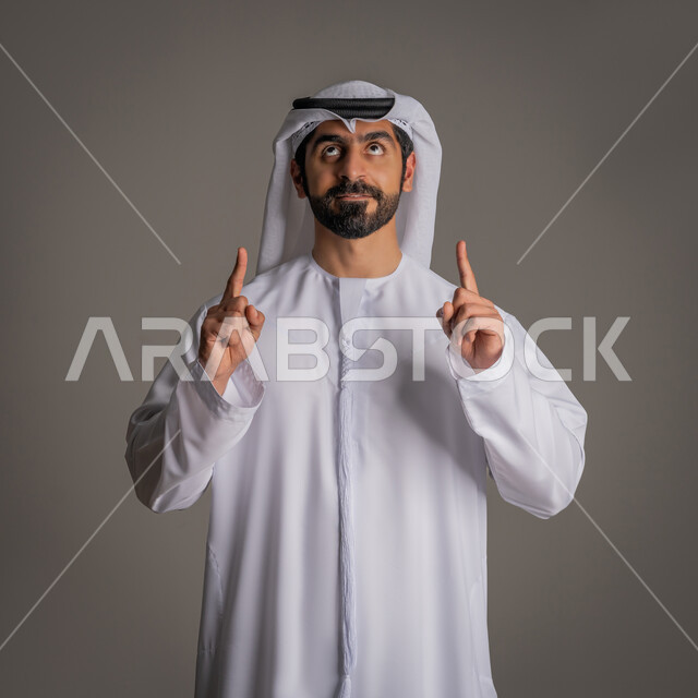 الوقوف باستقامة ورفع  اصبعي السبابة في كلتا اليدين, بورتريه لرجل عربي خليجي إماراتي يرتدي الكندورة البيضاء والغترة ينظر للأعلى بإيماءات الترجي والأمل, مفهوم الأناقة والرجولة والاهتمام بالمظهر الخارجي, خلفية رمادية