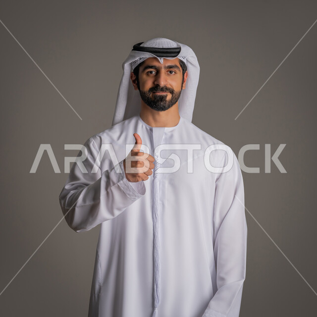 The concept of elegance, masculinity, and concern for external appearance, a portrait of an Emirati Gulf Arab man wearing a white kandora and ghutra, raising his thumb in a gesture of approval and acceptance, standing upright and looking at the camera with happy expressions, gray background