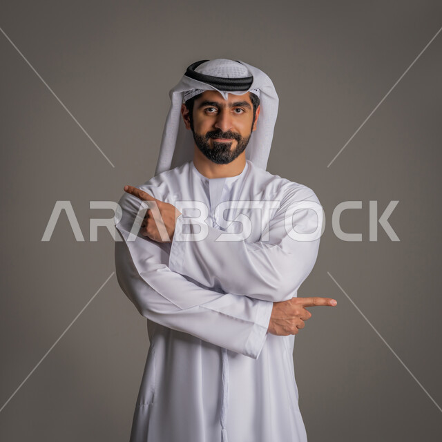 الوقوف باستقامة والنظر إلى الكاميرا بتعابير السعادة, بورتريه لرجل عربي خليجي إماراتي يرتدي الكندورة البيضاء والغترة, حركات اليدين والإشارة بأصابع السبابة إلى اتجاهين متعاكسين بإيماءات الثقة بالنفس, مفهوم الأناقة والرجولة والاهتمام بالمظهر الخارجي, خلفية رمادية