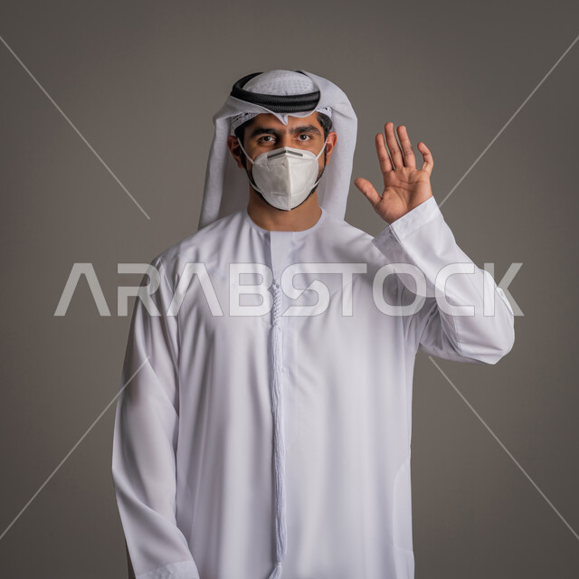 Raising the hand with a gesture to avoid shaking hands, be careful of the spread of the Corona virus, portrait of an Emirati Gulf Arab man wearing the Emirati kandora and ghutra and wearing a medical mask, caution against disease transmission, wearing a mask to stop Covid 20 infection, following safety procedures, gray background