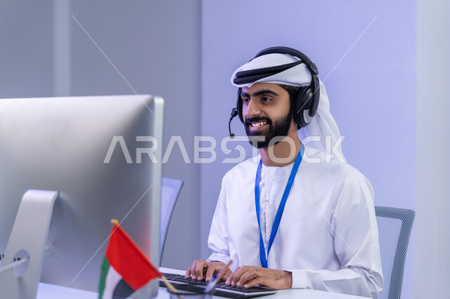 الإجابة على أسئلة العملاء، دمج التقنيات الحديثة في الحياة العملية، الفخر في الانتماء لدولة الإمارات، تعابير البهجة والسرور، بورتريه مقرب لموظف عربي خليجي اماراتي يرتدي الزي التقليدي يجلس في المكتب يعمل على جهاز الحاسوب، خلفية رمادية