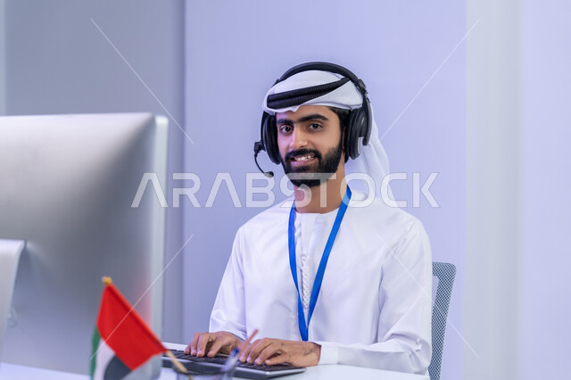 الفخر في الانتماء لدولة الإمارات، تعابير البهجة والسرور والنظر الى الكاميرة، دمج التقنيات الحديثة في الحياة العملية، بورتريه مقرب لموظف عربي خليجي اماراتي يرتدي الزي التقليدي يجلس في المكتب يعمل على جهاز الحاسوب، خلفية رمادية
