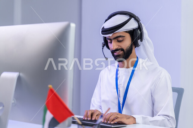 مفهوم العمل في مجال خدمة الزبائن، الفخر في الانتماء لدولة الإمارات، ايماءات البهجة والسرور، بورتريه مقرب لموظف عربي خليجي اماراتي يرتدي الزي التقليدي يجلس في المكتب يعمل على جهاز الحاسوب، دمج التقنيات الحديثة في الحياة العملية، خلفية رمادية