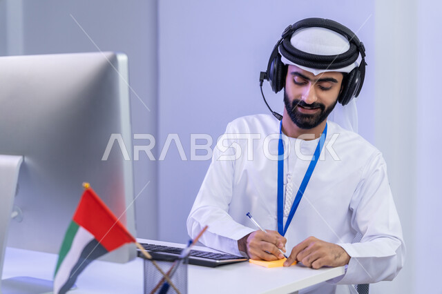 تعابير البهجة و السرور ، الفخر في الانتماء لدولة الإمارات، استخدام الأجهزة الحديثة، بورتريه مقرب لموظف عربي خليجي اماراتي يرتدي الزي التقليدي يجلس في المكتب يقوم بتدوين الملاحظات على الاوراق الملونة، دمج التقنيات الحديثة في الحياة العملية، خلفية رمادية