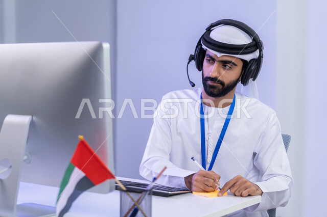الفخر في الانتماء لدولة الإمارات، ايماءات البهجة والسرور ، بورتريه مقرب لموظف عربي خليجي اماراتي يرتدي الزي التقليدي يجلس في المكتب و يقوم بتدوين الملاحظات على الورق الملون، دمج التقنيات الحديثة في الحياة العملية، خلفية رمادية