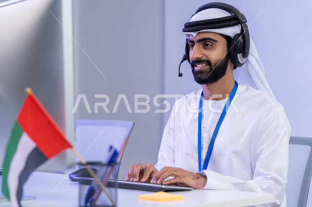 ايماءات البهجة والسرور، الاعتزاز والفخر في الانتماء لدولة الإمارات، دمج التقنيات الحديثة في الحياة العملية، بورتريه مقرب لموظف عربي خليجي اماراتي يرتدي الزي التقليدي يجلس في المكتب يعمل على جهاز الحاسوب بتعابير السرور، خلفية رمادية