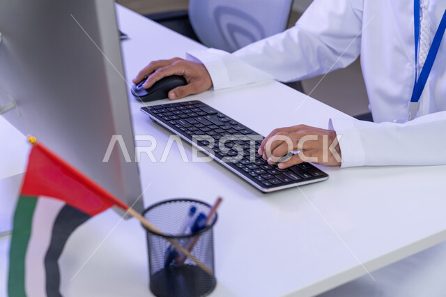 مفهوم العمل في مجال خدمة الزبائن، الاعتزاز في الانتماء لدولة الإمارات، اعداد وتقديم البرامج الإذاعية، الإجابة على أسئلة العملاء، بورتريه مقرب لأيدي موظف عربي خليجي اماراتي يرتدي الزي التقليدي يجلس في المكتب يعمل على جهاز الحاسوب