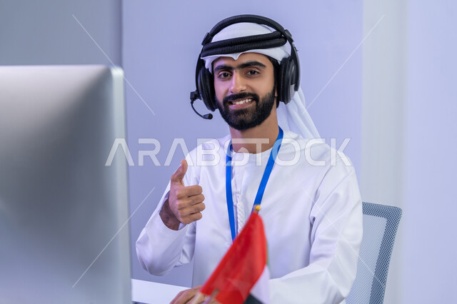 Gestures of joy and happiness, preparing and presenting radio programs, the concept of working in the field of customer service, a close-up portrait of an Arab Gulf Emirati employee wearing a ghutra and a gandoura sitting in the office raising his thumb up with expressions of satisfaction and admiration and a quality mark, gray background