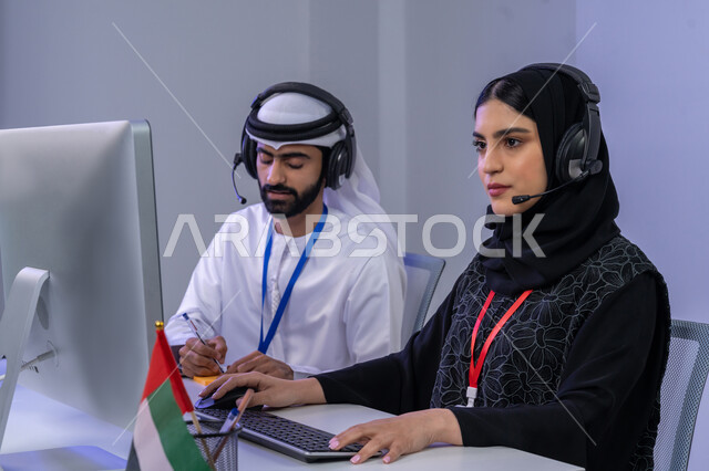 الاعتزاز والفخر في الانتماء لدولة الإمارات، ايماءات البهجة والسرور، استخدام الأجهزة التقنية الحديثة، بورتريه مقرب لموظفيين عربيين خليجيين اماراتيين يجلسان في المكتب يقومان بالتعاون والتشارك في اداء المهام، مساهمة النساء في الوظائف المكتبية، خلفية رمادية