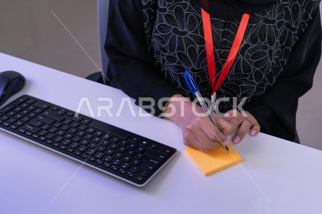 استخدام الأجهزة الحديثة، الإجابة على أسئلة العملاء، مساهمةالنساء في الوظائف المكتبية، مفهوم العمل في مجال خدمة الزبائن، بورتريه مقرب لأيدي موظفة عربية خليجية اماراتية تجلس في المكتب تقوم بتدوين الملاحظات على ملصقات الورق الملون، القيام بأداء المهام اليومية