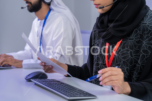مشاركة النساء في الوظائف المكتبية، مفهوم العمل في مجال خدمة الزبائن، استخدام الأجهزة التقنية الحديثة، الإجابة على أسئلة العملاء، بورتريه مقرب لموظفان عربيين خليجيين اماراتيين يجلسان في المكتب يقومان بالتعاون والتشارك في اداء المهام، خلفية رمادية
