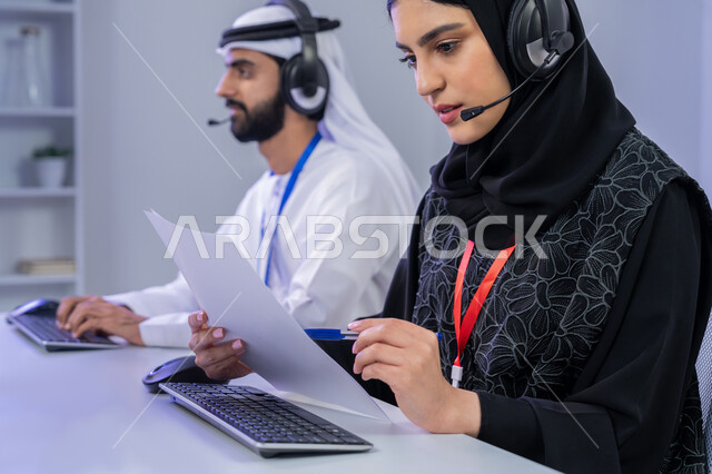 Answering customer questions, using modern technical devices, concept of working in the field of customer service, close-up portrait of two Emirati Gulf Arab employees sitting in the office cooperating and sharing tasks, women participating in office jobs, gray background