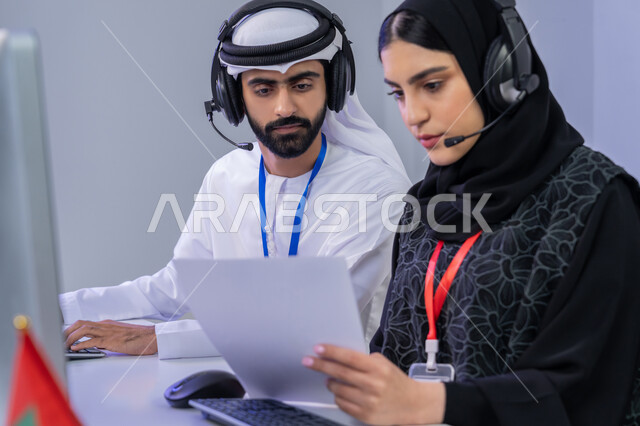 Using modern technology devices, answering customer questions, customer service work concept, close-up portrait of two Emirati Gulf Arab employees sitting in the office cooperating and sharing tasks, women participating in office jobs, gray background