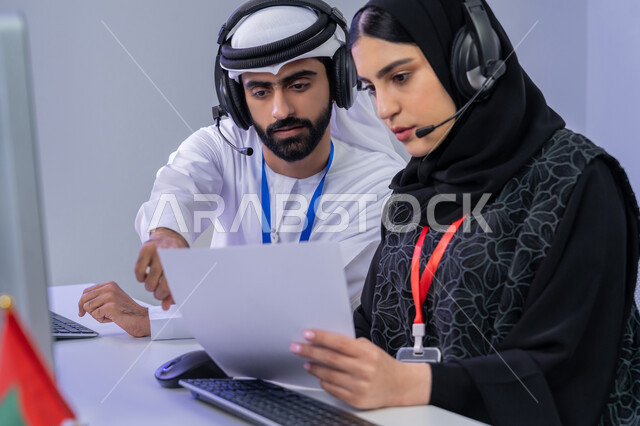 مفهوم العمل في مجال خدمة الزبائن، استخدام الأجهزة التقنية الحديثة، الإجابة على أسئلة العملاء، بورتريه مقرب لموظفان عربيين خليجيين اماراتيين يجلسان في المكتب يقومان بالتعاون والتشارك في اداء المهام، مشاركة النساء في الوظائف المكتبية، خلفية رمادية